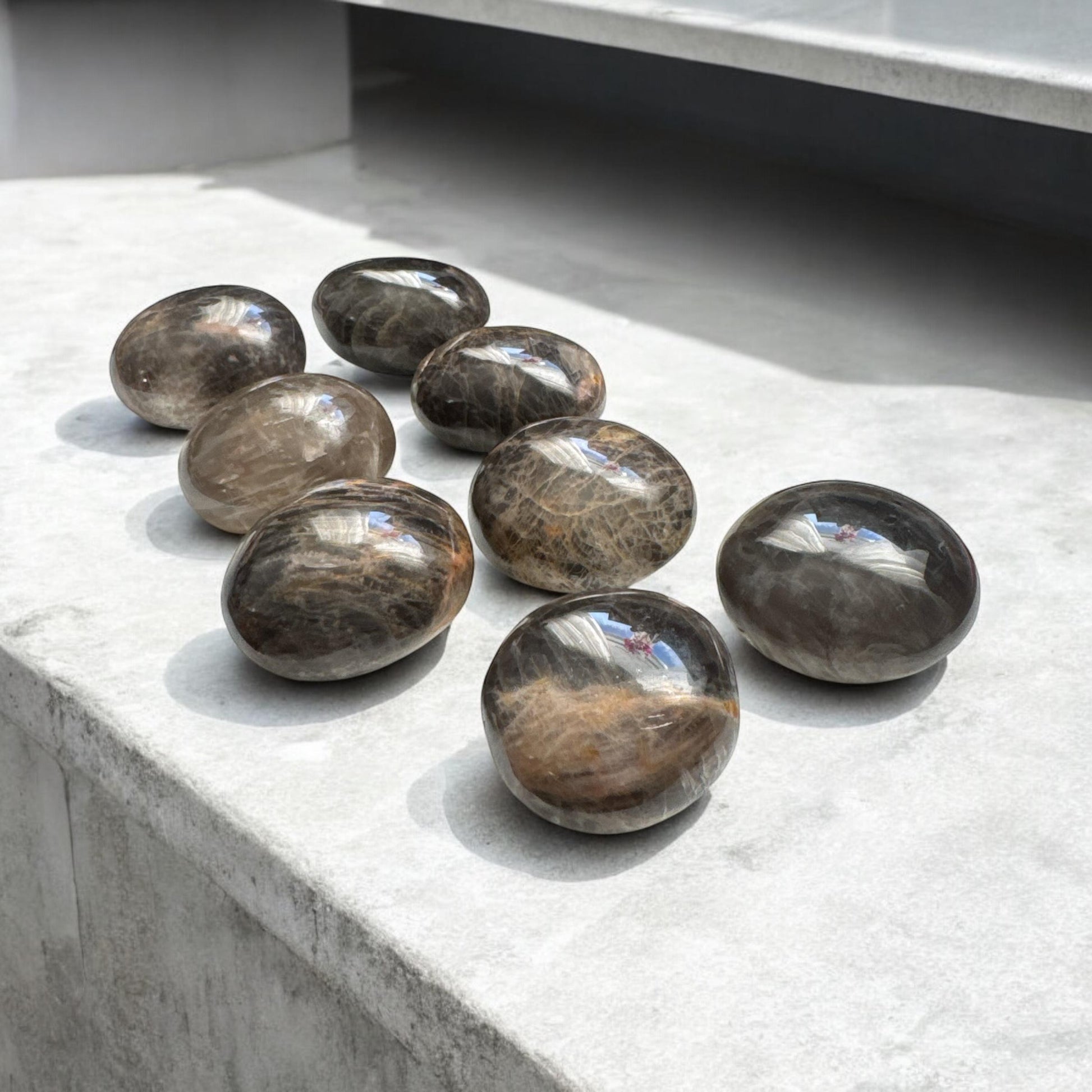 May include: Eight smooth, black and brown gemstone spheres, also known as black moonstone, arranged in a row on a light grey surface.