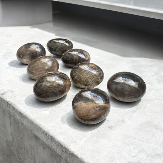 May include: Eight smooth, black and brown gemstone spheres, also known as black moonstone, arranged in a row on a light grey surface.