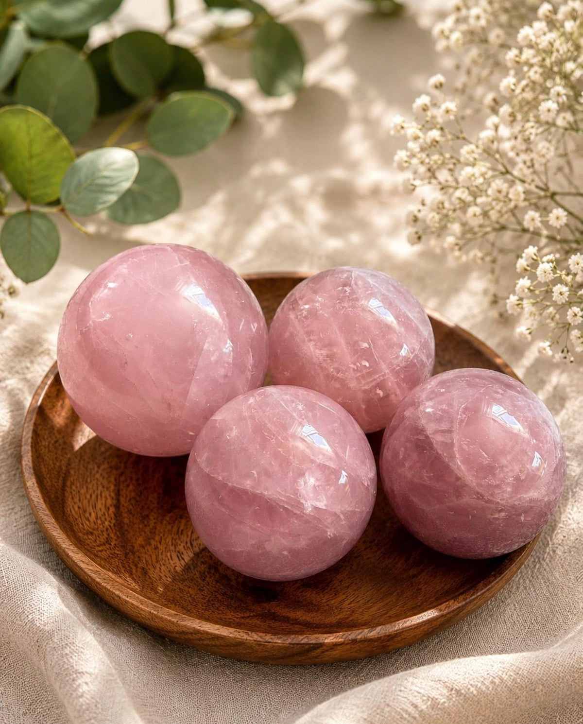 May include: Four polished rose quartz spheres of varying sizes arranged on a circular wooden tray. The stones are translucent, light pink in colour, and feature natural internal fractures and crystalline textures throughout their smooth surfaces.